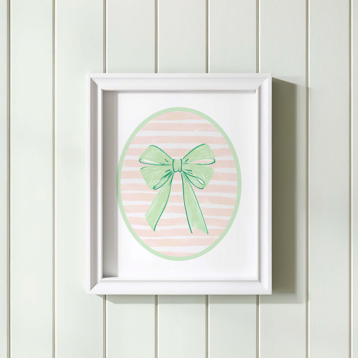 Green bow nursery wall art print with a pink and white striped oval background, shown in a wooden frame. Chic and preppy decor for a baby girl's room.