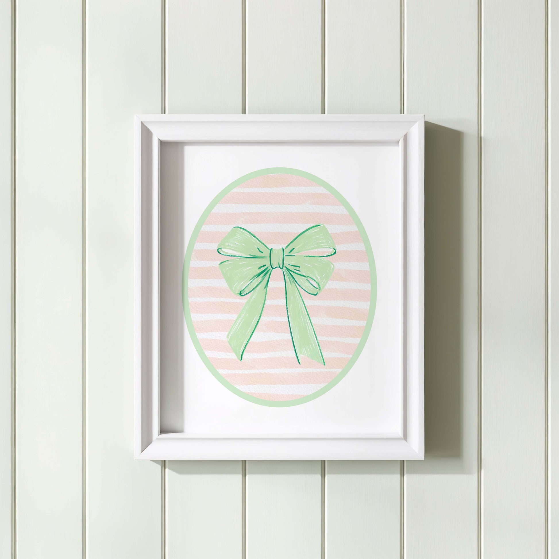 Green bow nursery wall art print with a pink and white striped oval background, shown in a wooden frame. Chic and preppy decor for a baby girl's room.