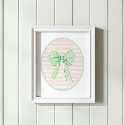Green bow nursery wall art print with a pink and white striped oval background, shown in a wooden frame. Chic and preppy decor for a baby girl's room.