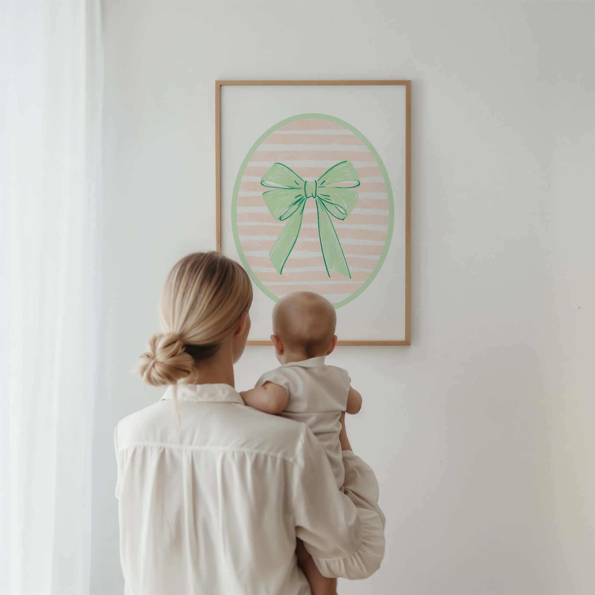 Green bow nursery wall art print with a pink and white striped oval background, shown in a wooden frame. Chic and preppy decor for a baby girl's room.
