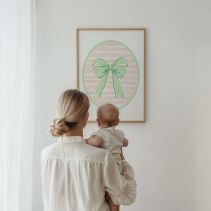 Green bow nursery wall art print with a pink and white striped oval background, shown in a wooden frame. Chic and preppy decor for a baby girl's room.