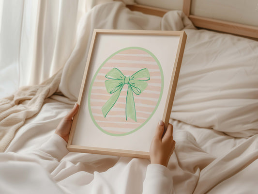 Green bow nursery wall art print with a pink and white striped oval background, shown in a wooden frame. Chic and preppy decor for a baby girl's room.